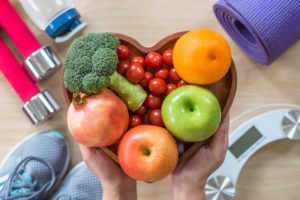 a person holding a heart-shaped bowl of healthy foods. in the background is a weight scale, yoga mat, water bottle, tennis shoes, and weights